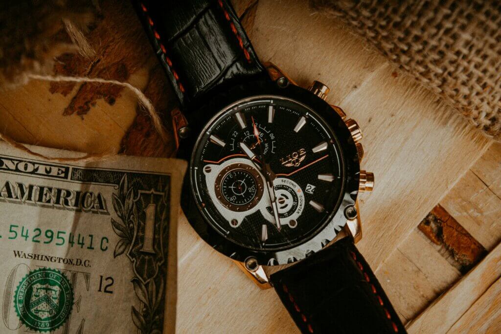 A premium leather watch with an elaborate clock face rests on a piece of wood, with a dollar bill in the bottom left corner.
